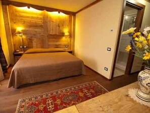 A bedroom with a wooden headboard, a bed, a nightstand, a bedside table, a vase with flowers, and a patterned rug. - Hotel Mon Reve (Valtournenche)