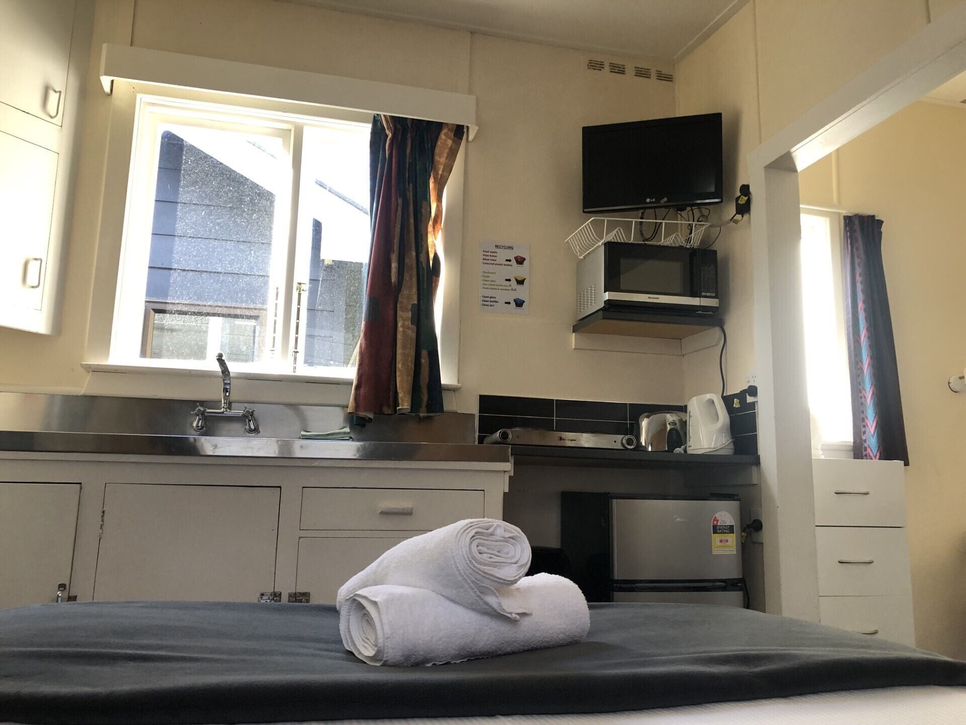A hotel room with a bed, a window with curtains, a kitchenette with a microwave, a kettle, and a TV on the wall.