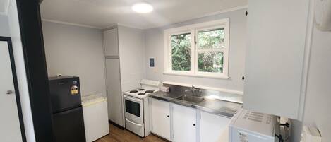 Cocina compacta con armarios blancos, estufa, refrigerador y ventana con vistas a la vegetación.
