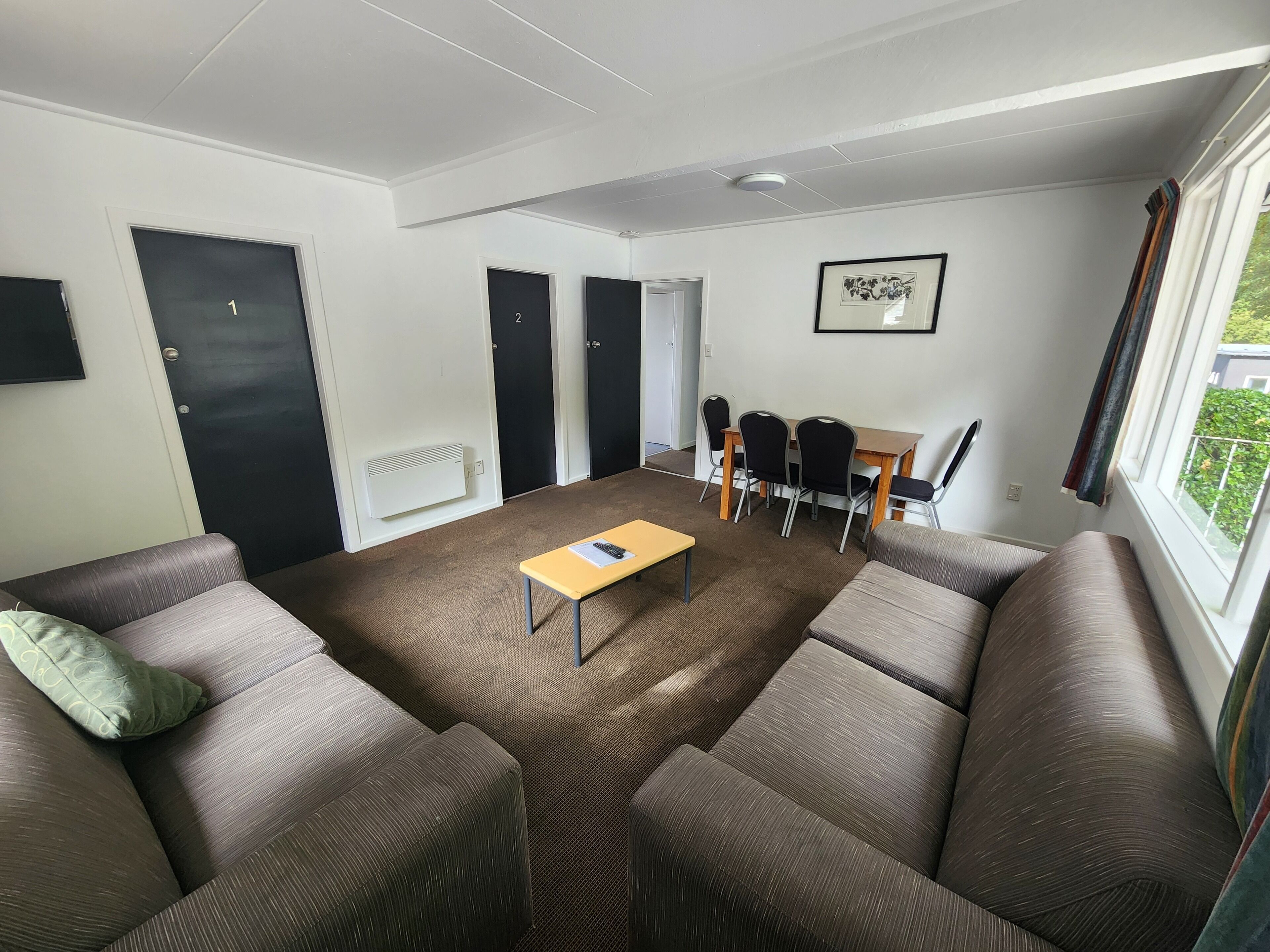 Ein modernes Wohnzimmer mit einem braunen Ecksofa, einem kleinen gelben Tisch und einem Essbereich mit vier Stühlen. Es gibt zwei schwarze Türen, eine weiße Heizung und ein gerahmtes Bild an der Wand.