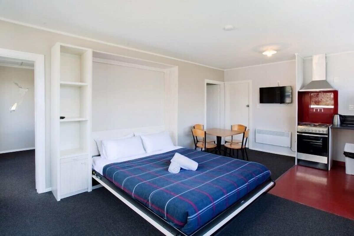 A compact hotel room with a bed, a small kitchenette, a dining area, and a TV.