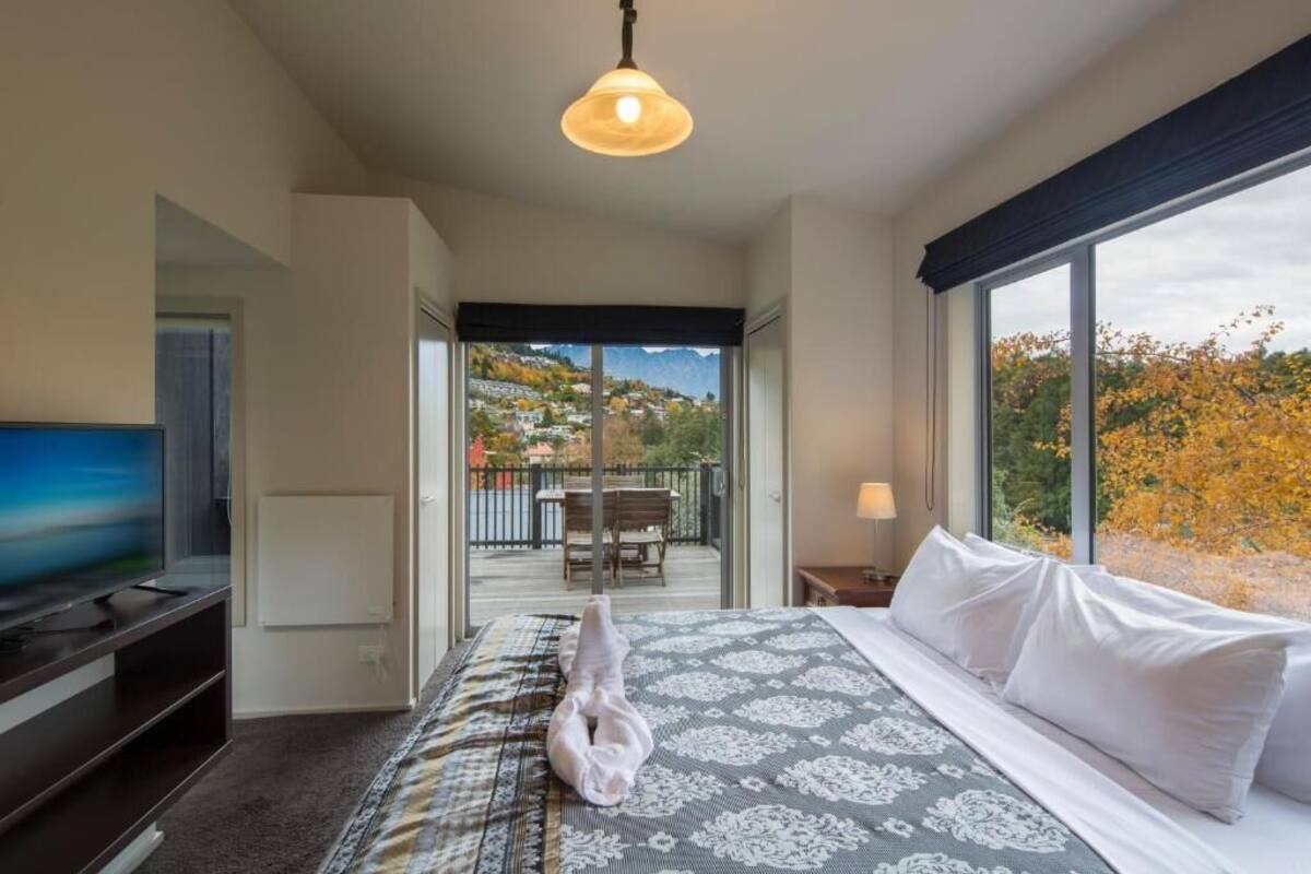 A hotel room with a bed, a TV, a balcony with a view of a hillside, and a hanging light fixture.