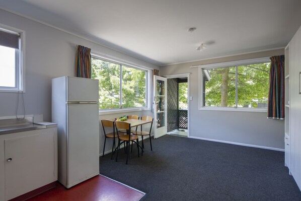 A compact kitchen with a refrigerator, a dining table with chairs, and large windows offering views of trees. - Pinewood Queenstown (Queenstown)