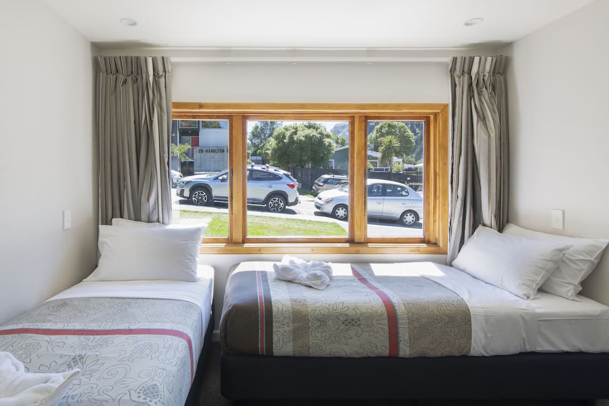 A hotel room with two beds, a large window, and a view of parked cars outside.