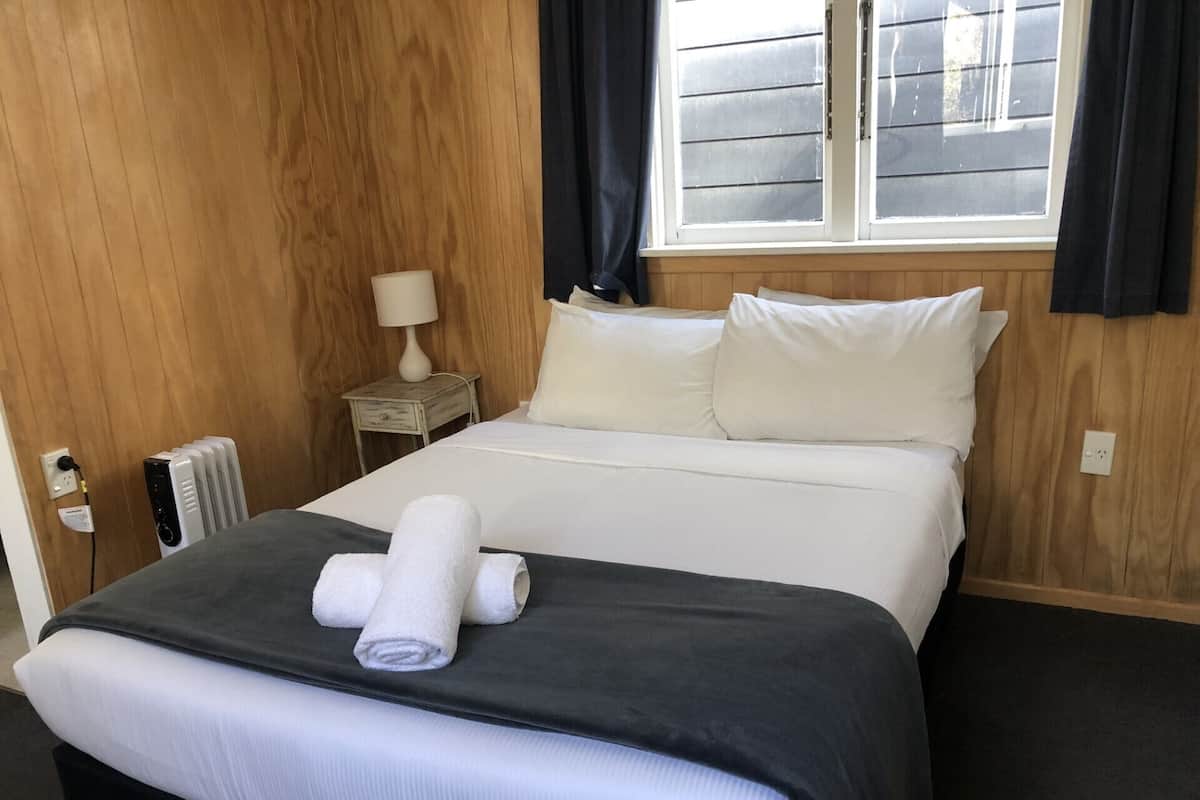 A hotel room with a bed, a bedside table with a lamp, a window with curtains, and a radiator.