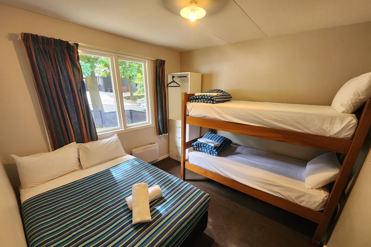 A room with a single bed, a bunk bed, a radiator, a window with curtains, and a hanging coat rack.
