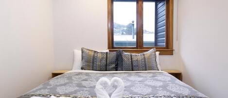 Una cama bien tendida con una disposición de toallas en forma de corazón, una ventana con persianas y vistas a un paisaje nevado.