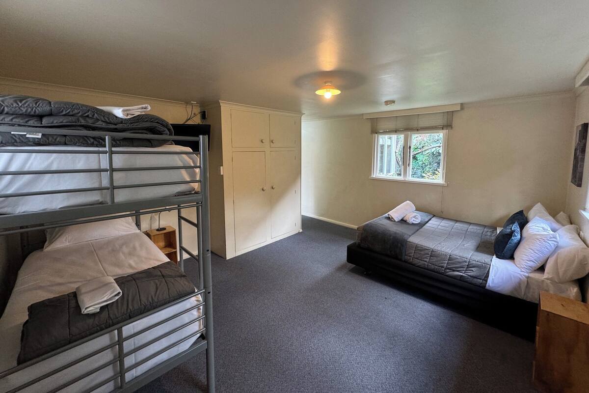 A dormitory room with bunk beds, a single bed, a wardrobe, and a window with a view of greenery.
