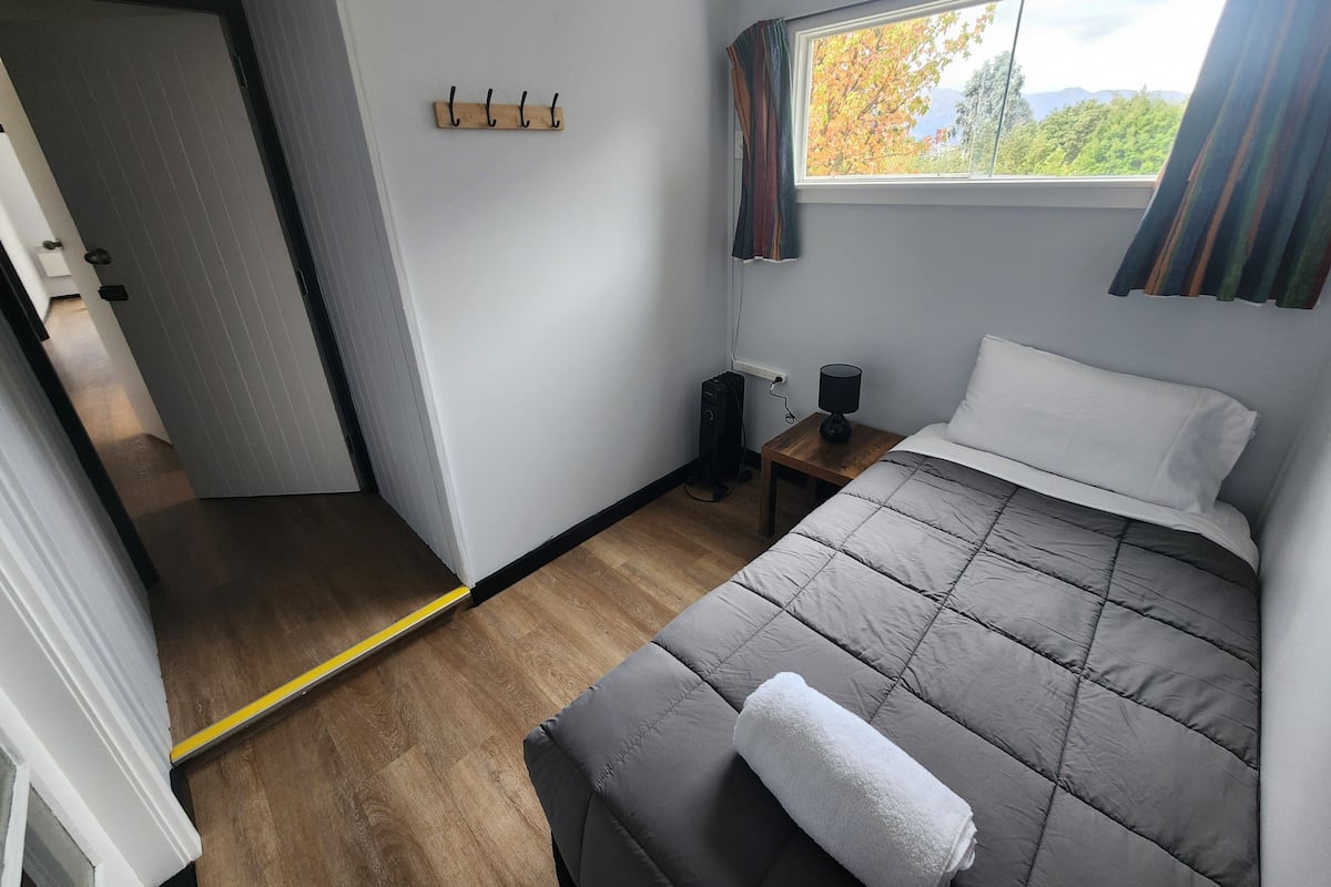 A bedroom with a bed, a bedside table with a lamp, a window with curtains, and a coat rack.