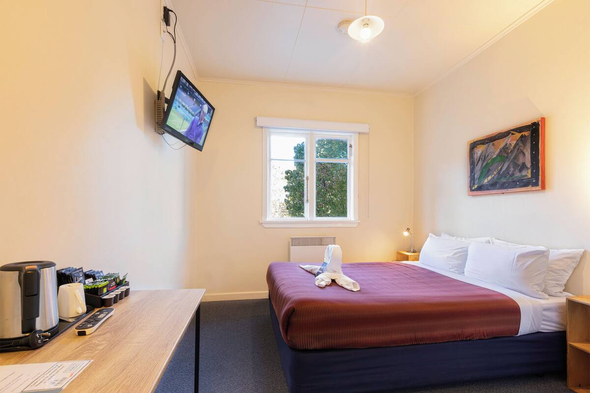 A hotel room with a bed, a desk with a coffee maker, a television, and a window with a view of trees.