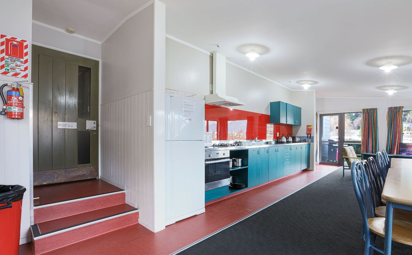 Een moderne keuken met turquoise en witte kastjes, een rode aanrecht en een zwarte mat die leidt naar een eetgedeelte met blauwe stoelen.