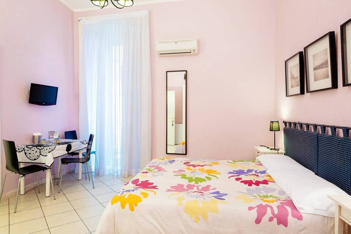 A bedroom with a bed, a desk, and a chair. There is a TV mounted on the wall and an air conditioning unit.