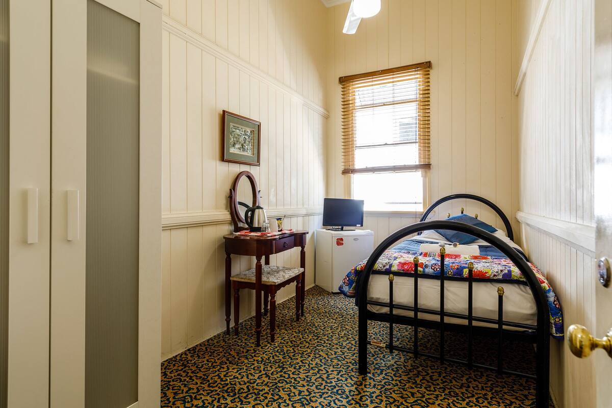 A bedroom with a bed, a desk with a chair, a television, and a window with blinds.