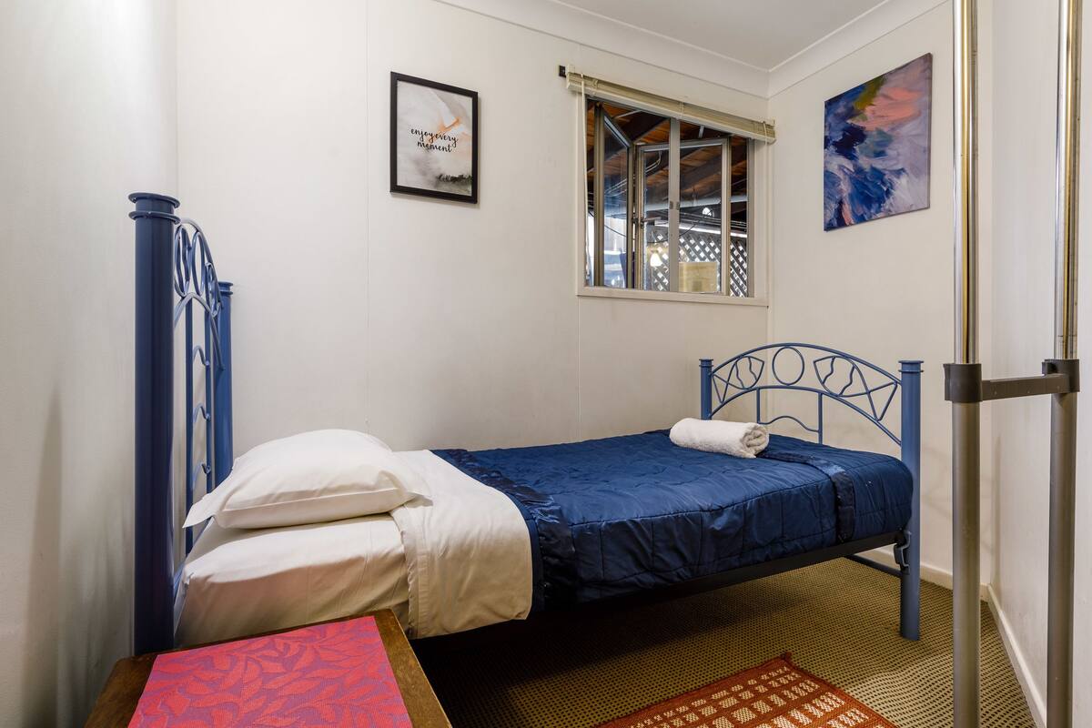 A single bed with a blue headboard and white bedding, a small window with a view, and a framed picture on the wall.