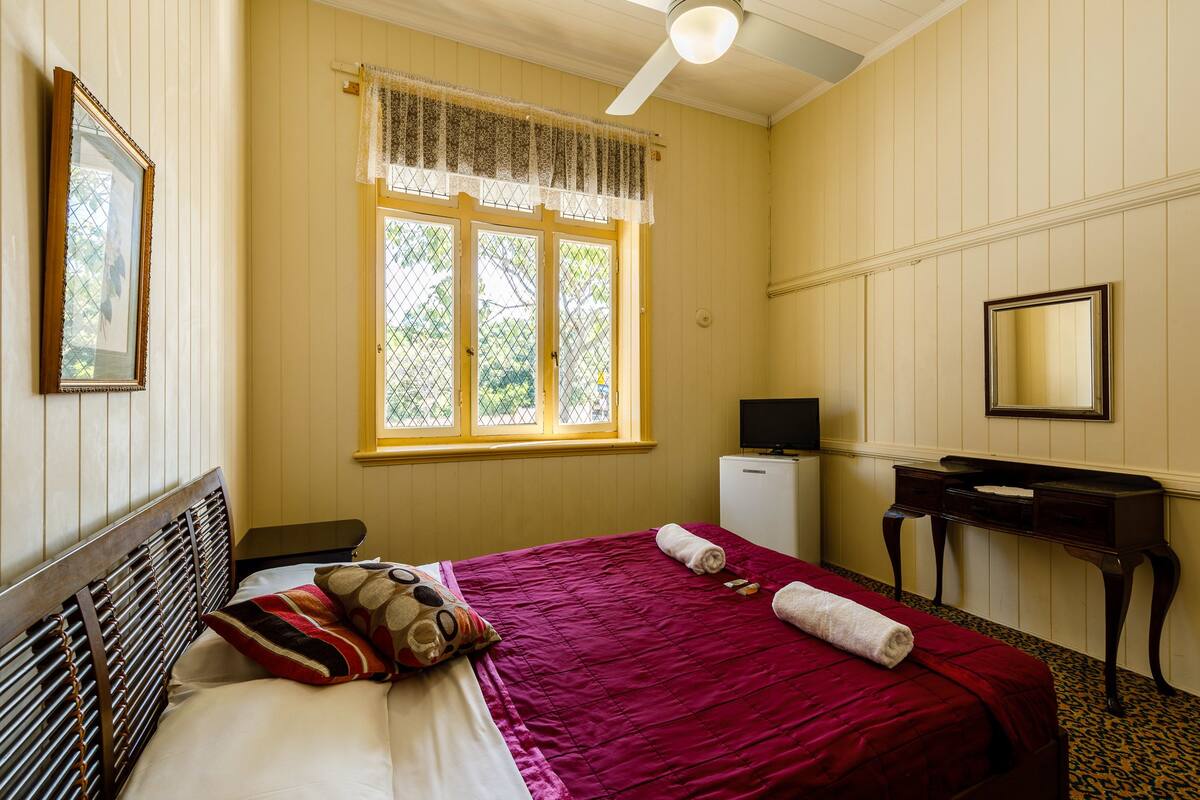 A bedroom with a bed, a desk, a chair, a television, and a window with a lace curtain.
