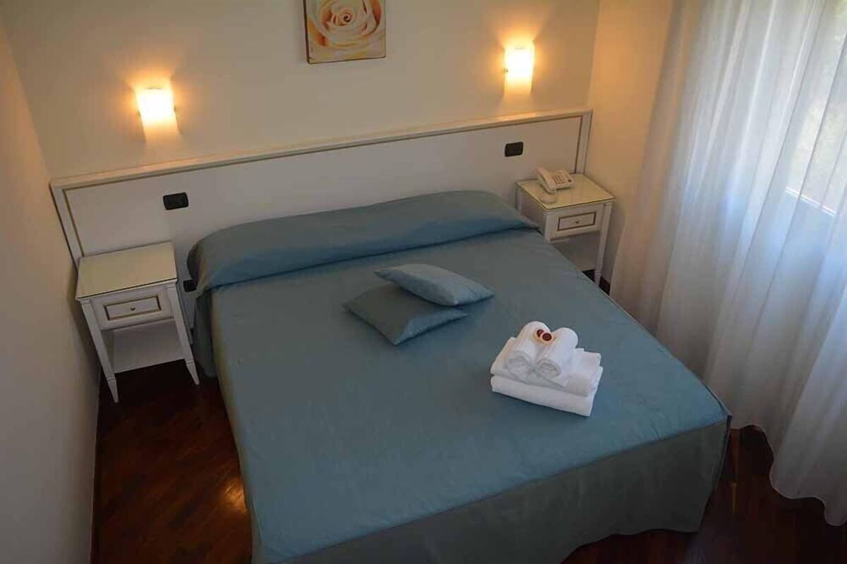 A neatly made bed with a blue bedspread, two pillows, and a folded white towel on the bed. There are two bedside tables, each with a lamp and a telephone.