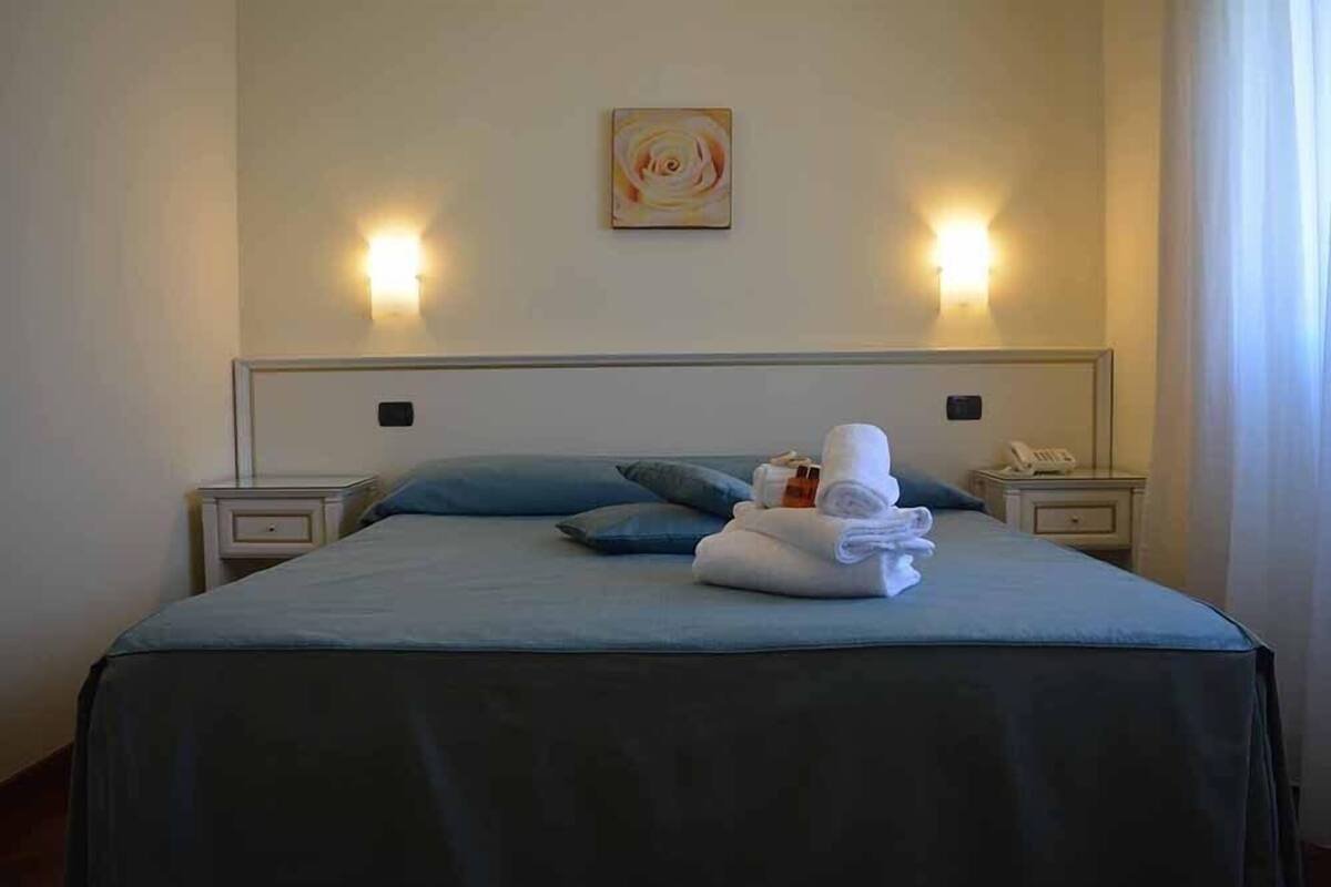A neatly made bed with a blue bedspread, two pillows, and a stack of folded towels. There are two bedside tables with lamps and a telephone.
