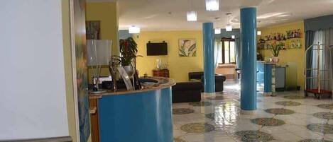 Un vestíbulo con una mesa de recepción, pilares azules, suelo de baldosas y una zona de bar con botellas.