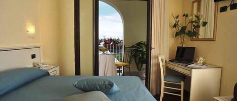 A hotel room with a bed, a desk with a laptop, a chair, a small table with a vase, and a view of the outdoors.