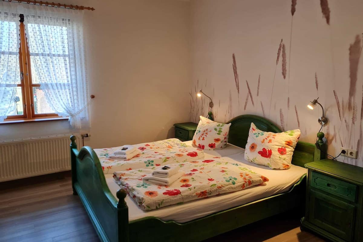 A bedroom with a green bed, floral bedding, a window with sheer curtains, and a wall with a rustic design.