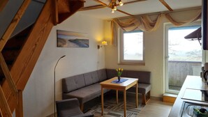 A cozy room with a sofa, a small table, a chair, a painting, a lamp, a window with curtains, and a staircase. - Reethäuser auf Rügen (Mönchgut-Granitz)