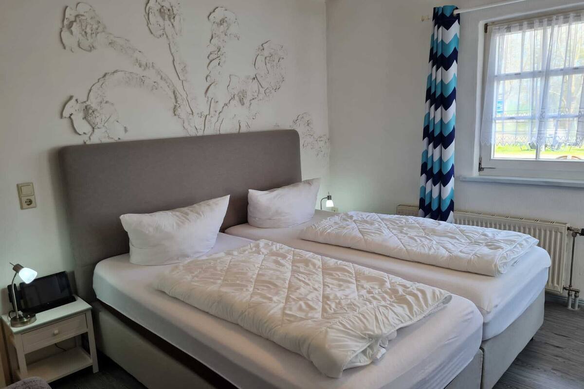 A double bed with a grey headboard, white bedding, a bedside table with a lamp, and a wall with a floral mural.