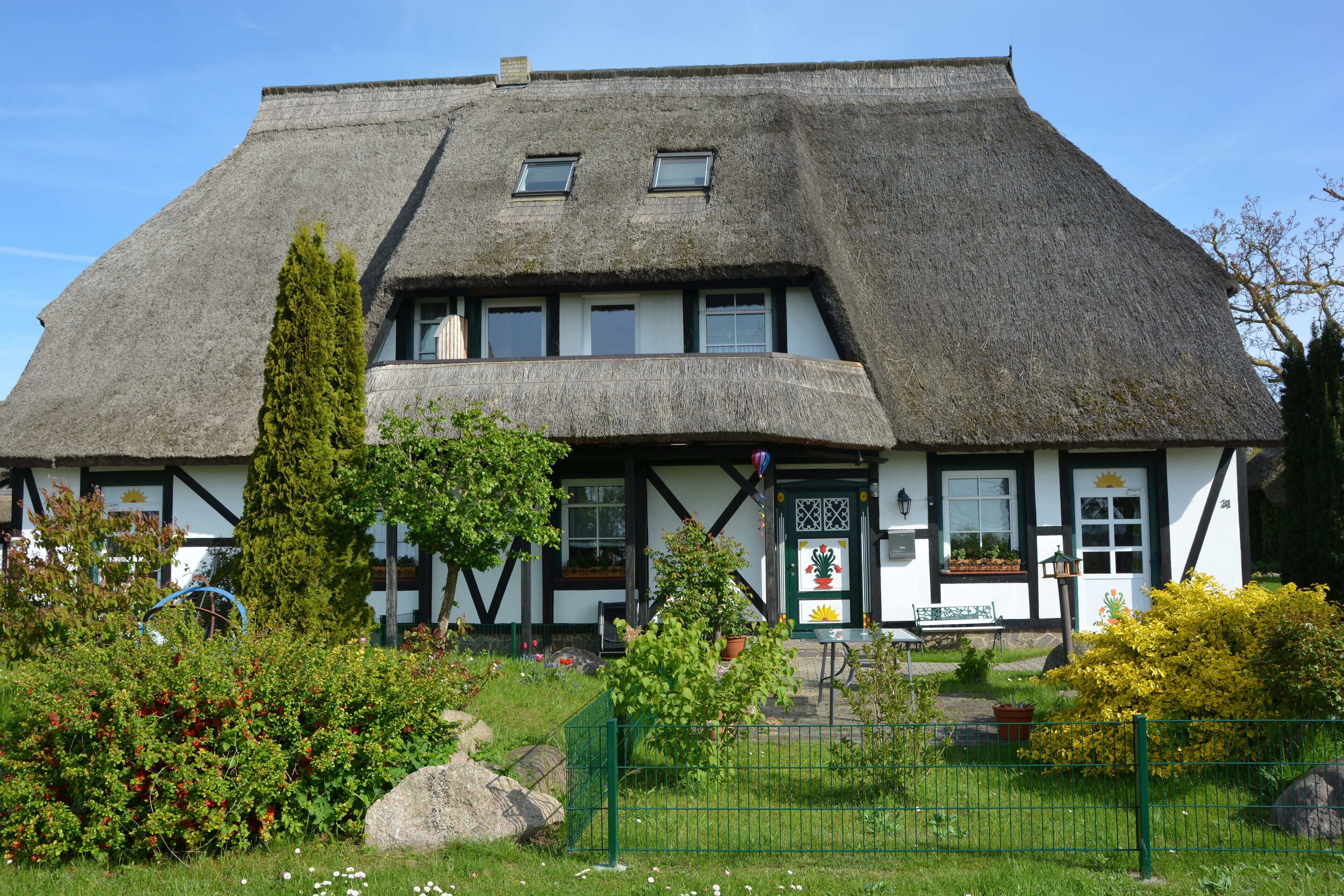A traditional thatched-roof building with a white exterior and black trim, surrounded by a well-maintained garden with various plants and a green fence.