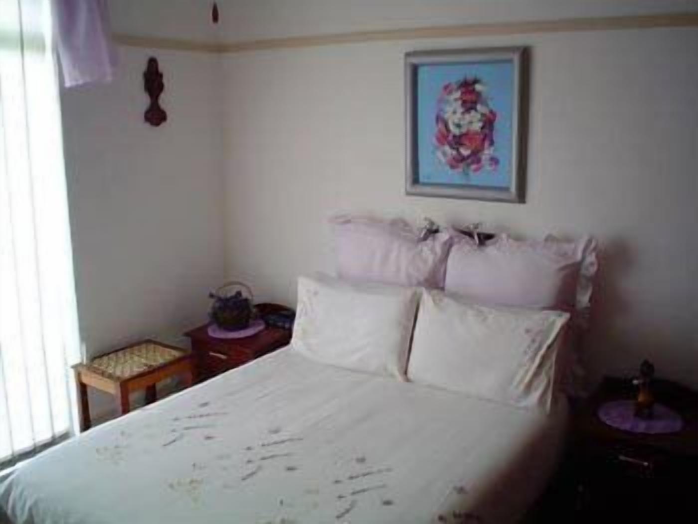 Un dormitorio con cama, almohadas, una mesita de noche, una pequeña mesa con lámpara y un cuadro en la pared.