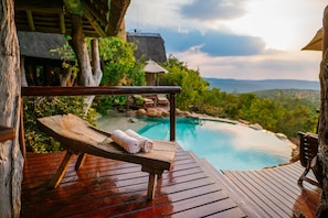 Terraza de madera con una silla de descanso y una toalla, con vistas a una piscina y un paisaje montañoso.