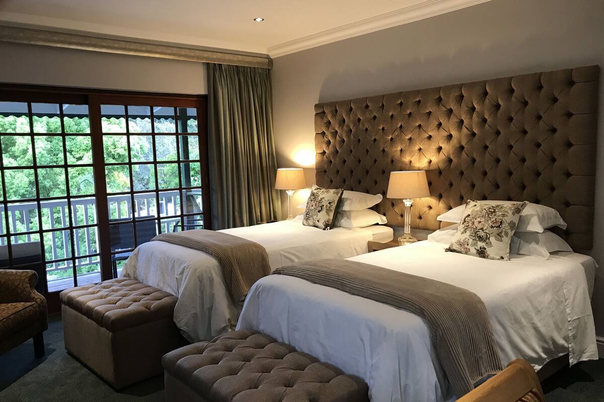 A hotel room with two beds, a large window with a view of greenery, a tufted headboard, and a wooden bench.