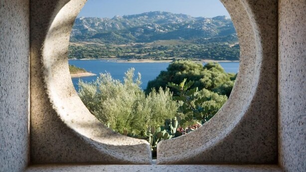 A circular window with a view of a lake, mountains, and greenery. - Liscia Country Relais (Luras)