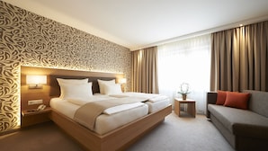 A hotel room with a large bed, a bedside table with a lamp, a small side table with a plant, a sofa with cushions, and a patterned wallpaper. - Hotel Hanauerhof (Appenweier)