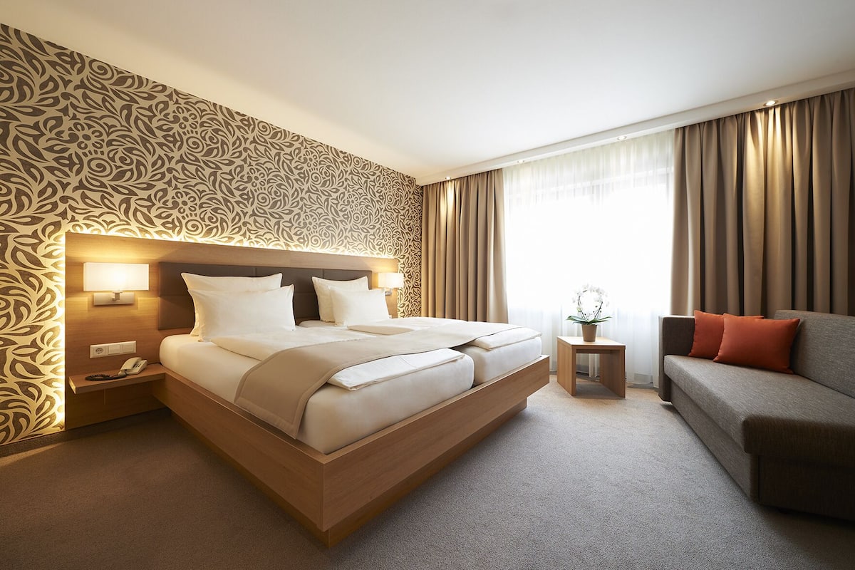 A hotel room with a large bed, a bedside table with a lamp, a small side table with a plant, a sofa with cushions, and a patterned wallpaper.