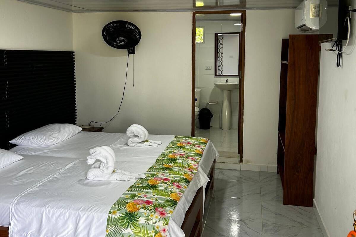 Habitación de hotel con cama, ventilador, armario y baño visible a través de una puerta abierta.