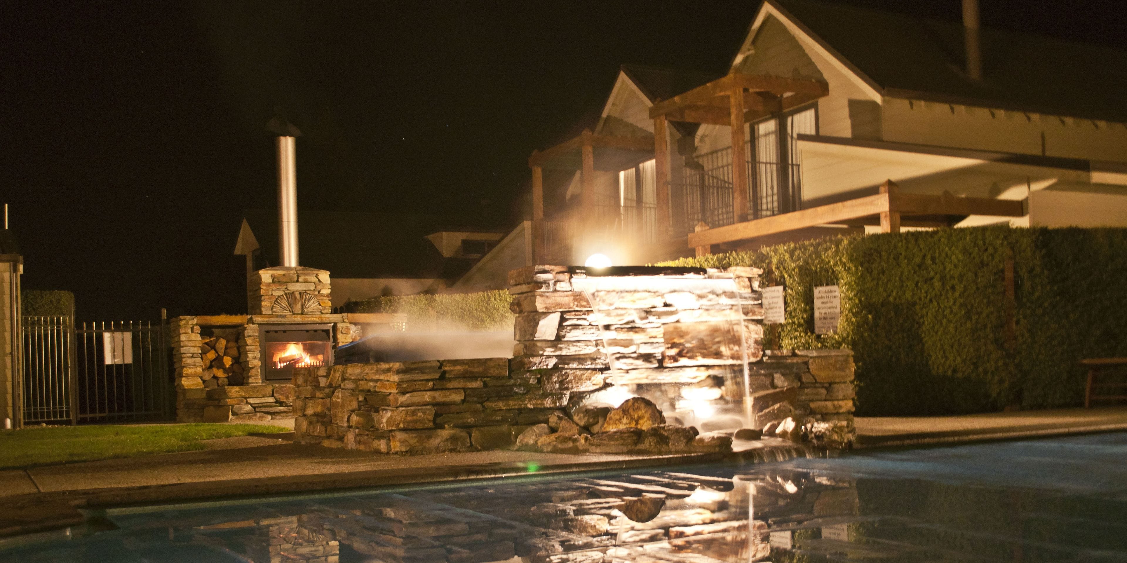 A stone structure with a fire feature and a water fountain, surrounded by a pool and a building with a balcony.