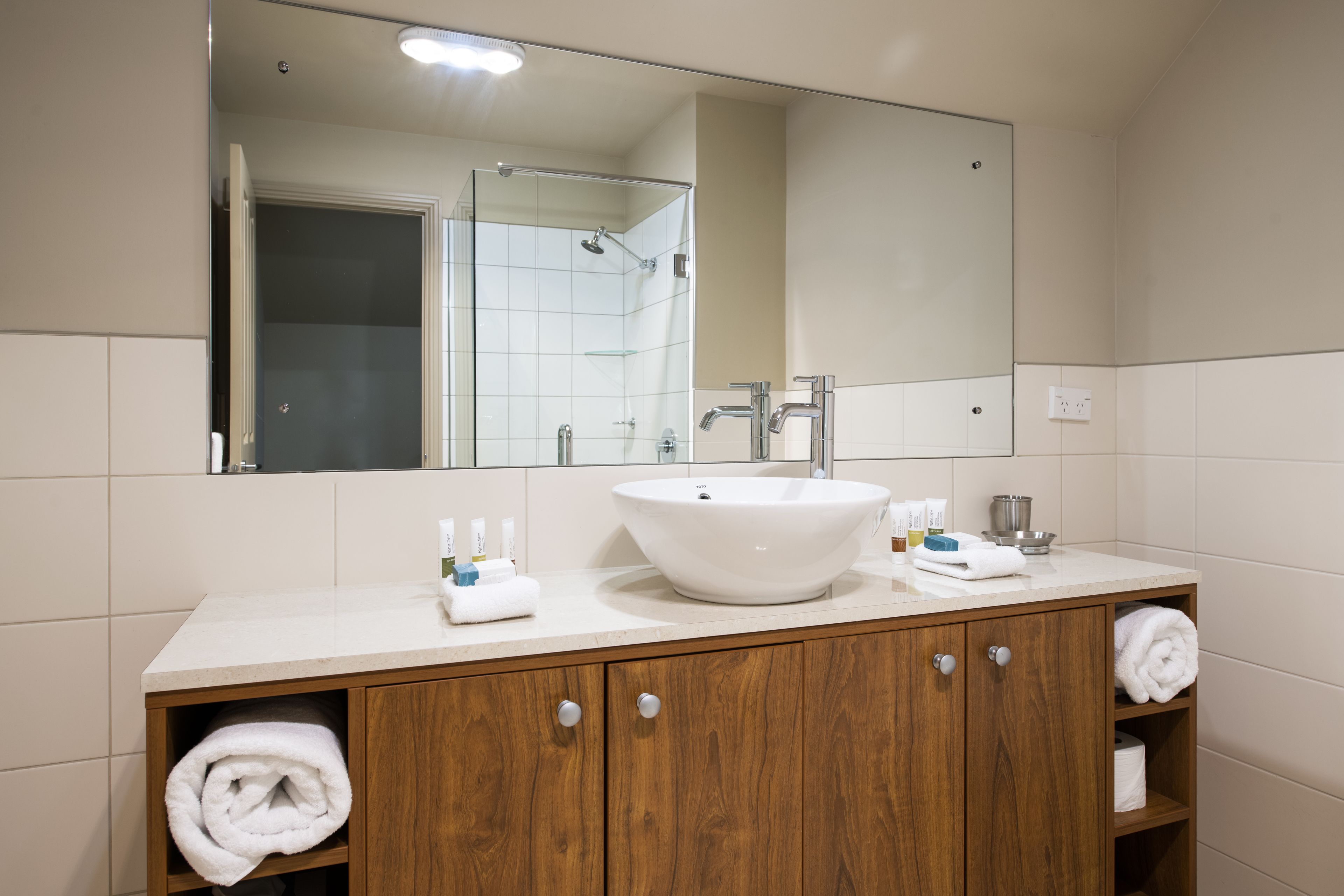 A bathroom with a large mirror, a white sink, wooden cabinets, and a shower area.
