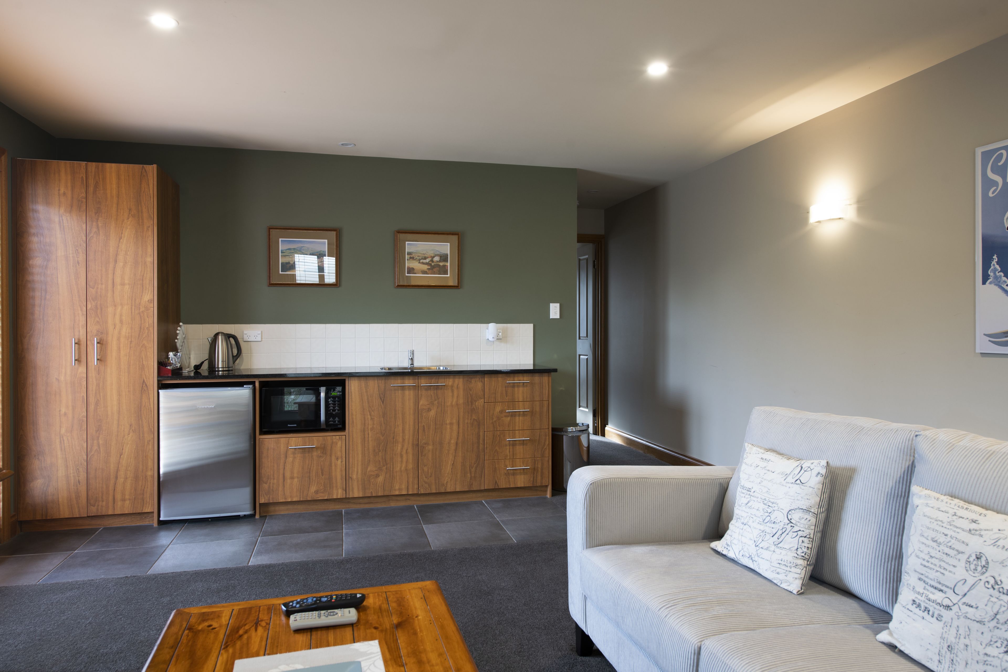 A modern kitchen with wooden cabinets, a stainless steel refrigerator, and a microwave. There is a sofa with decorative pillows and a coffee table with a remote control.