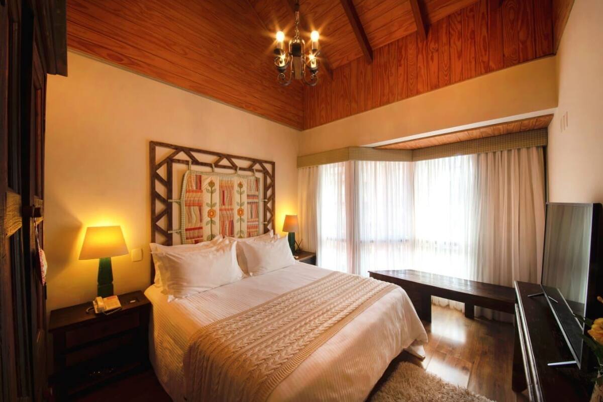 A hotel room with a wooden ceiling, a bed with white bedding, a nightstand with a lamp, a large window with curtains, and a flat-screen TV.