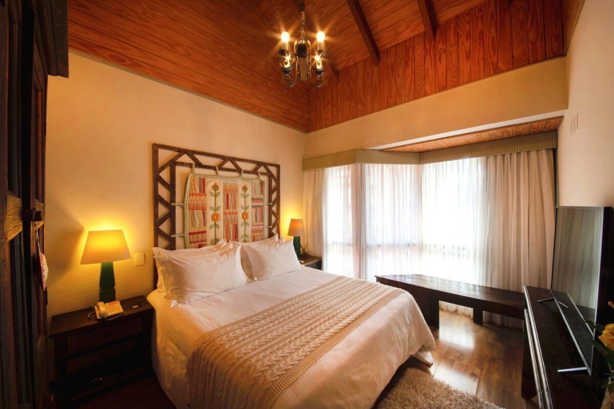 Habitación de hotel con techo de madera, cama con ropa de cama blanca, mesita de noche con lámpara, ventana grande con cortinas y televisor de pantalla plana.