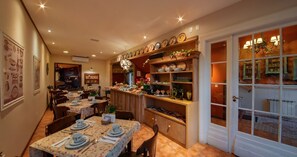 A dining area with tables set for a meal, a kitchen area with shelves of dishes, and a view through a glass door. - Hotel Petit Casa da Montanha (Gramado)
