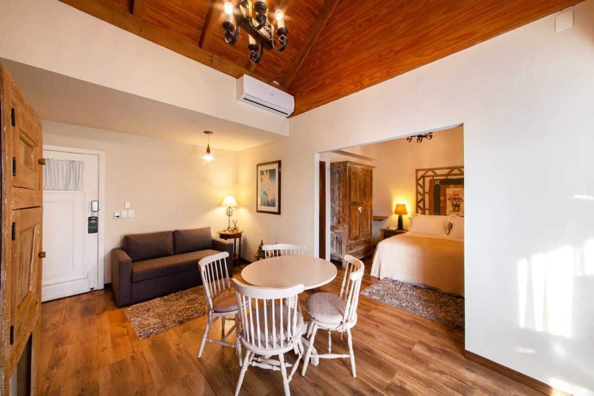 A cozy room with a wooden ceiling, a round dining table with chairs, a sofa, and a bed with a nightstand.