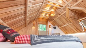 A cozy attic bedroom with wooden beams, a bed with patterned bedding, and a window with a view of greenery. - ALPIK Chalets - Bohinj (Bohinj)