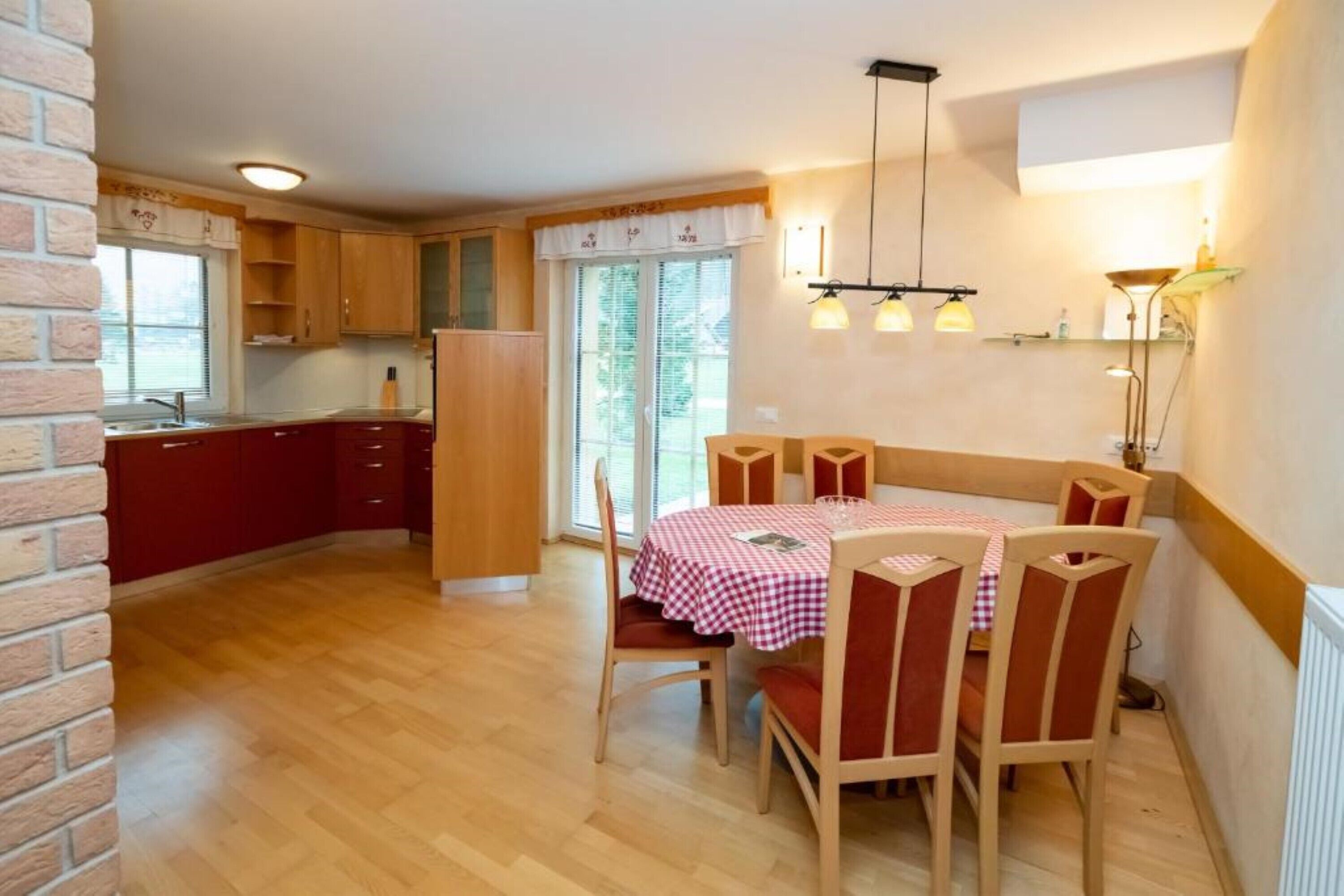 A compact kitchen with wooden cabinets, a red countertop, and a dining area with a checkered tablecloth and wooden chairs.