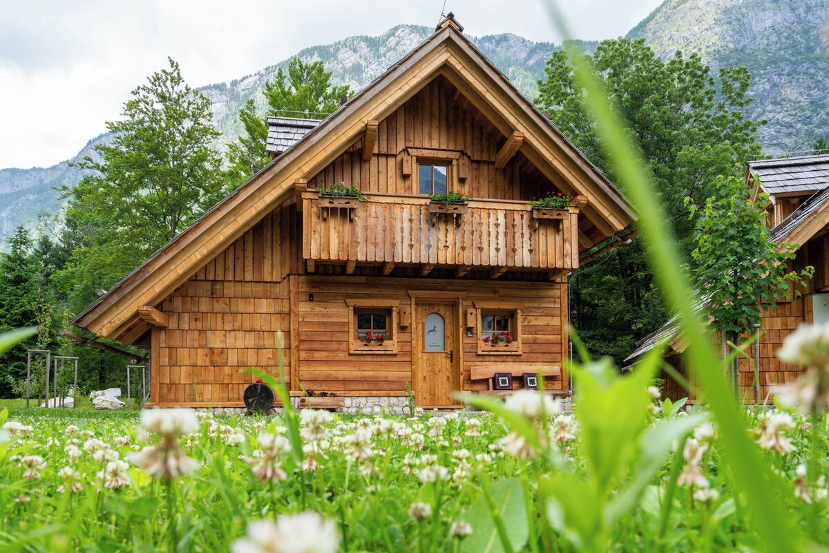 A wooden chalet with a balcony, surrounded by lush greenery and mountains.