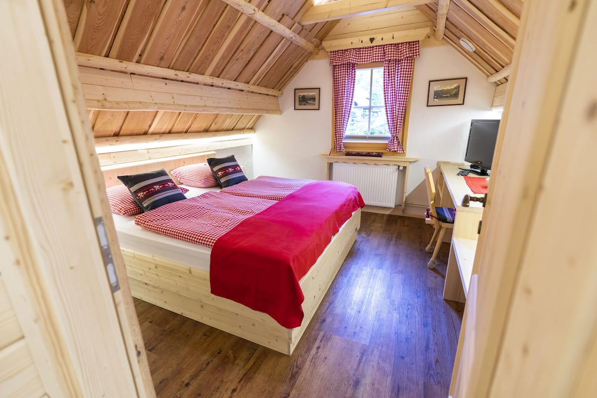 A cozy bedroom with a wooden ceiling, a bed with red bedding, a window with checkered curtains, and a desk with a computer.