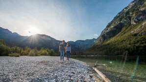A couple walking on a pebbled path beside a lake with mountains in the background. - ALPIK Chalets - Bohinj (Bohinj)