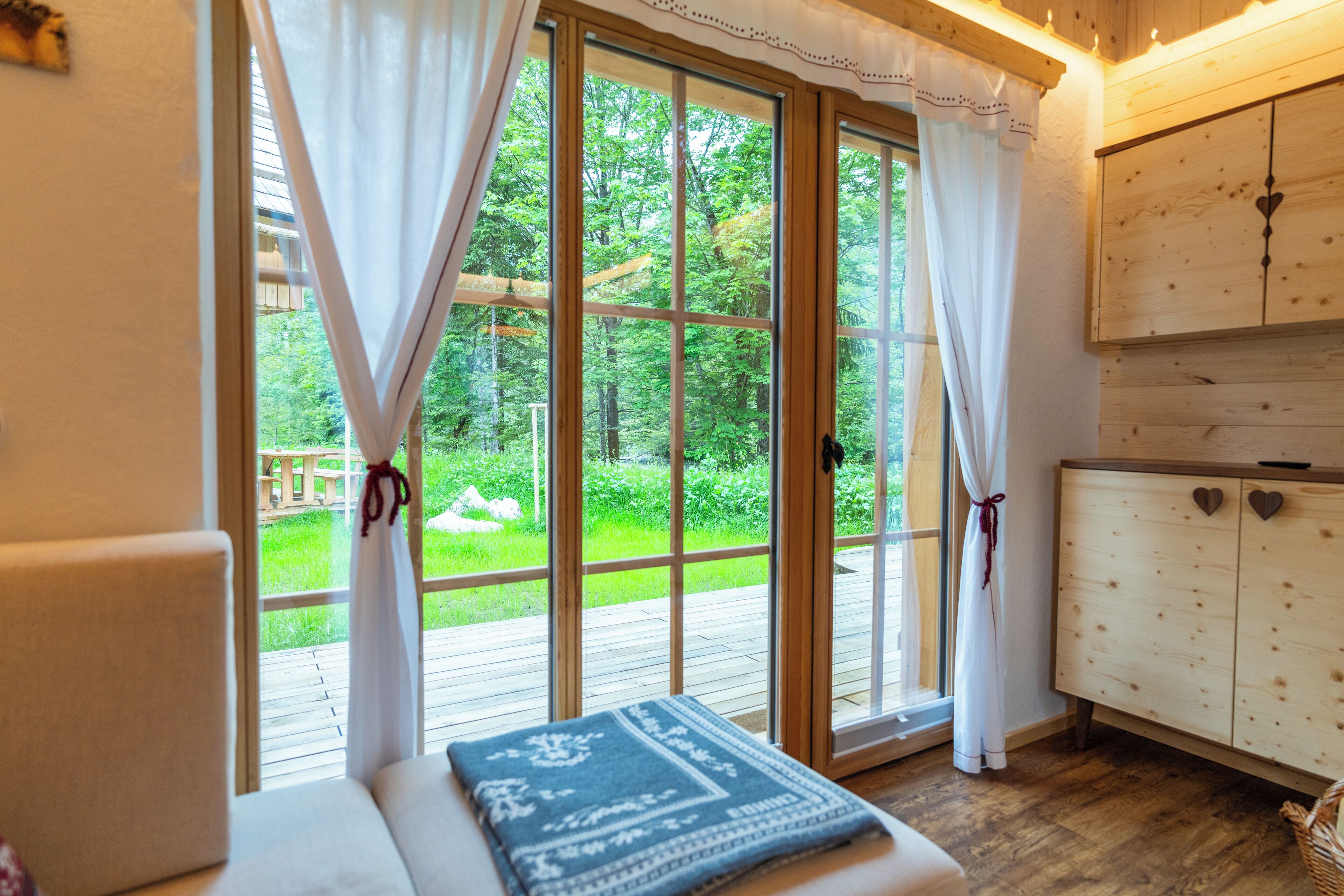 A room with a large sliding glass door leading to a wooden deck, a white cabinet with heart-shaped handles, and a bench with a patterned throw.