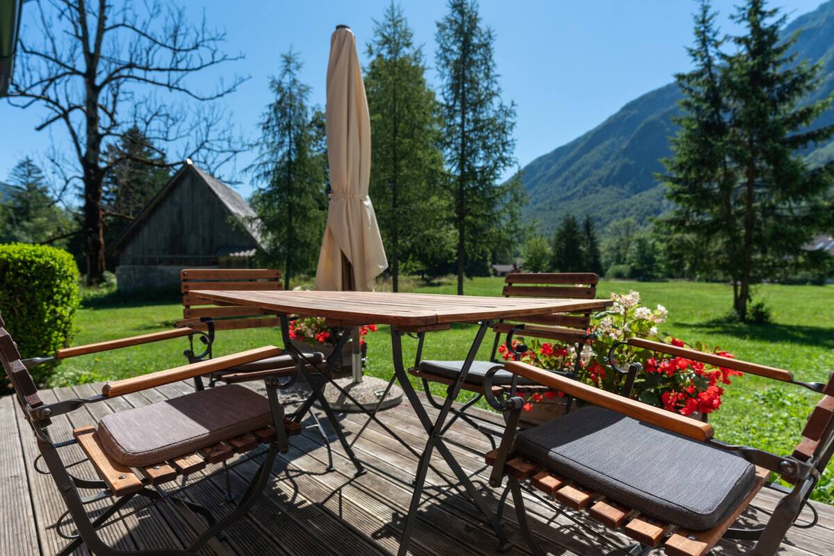 A wooden deck with a table and four chairs, surrounded by greenery and a mountain backdrop.