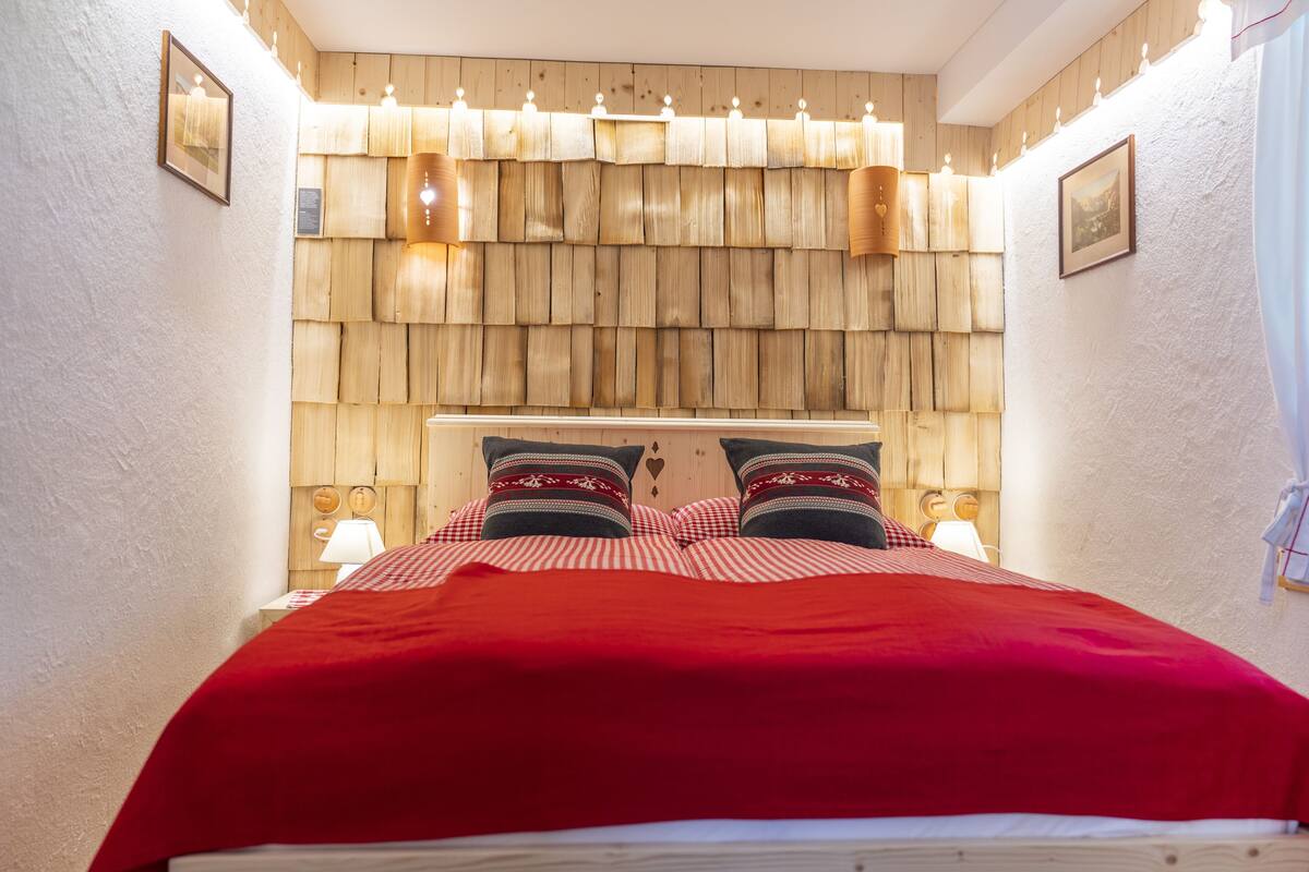 A neatly made bed with a red blanket and checkered pillowcases, wooden headboard with hanging lights, and two bedside lamps.