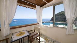 A balcony with a wooden ceiling, white curtains, a table with a sun design, and a view of the sea and a rocky coastline. - Hotel La Palma (Serrara Fontana)